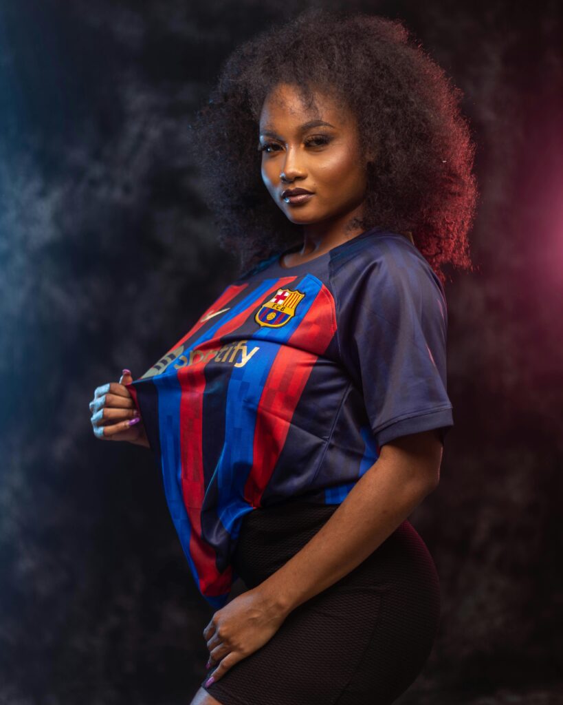 Confident woman with afro hairstyle wearing a Barcelona soccer jersey in a studio setting.